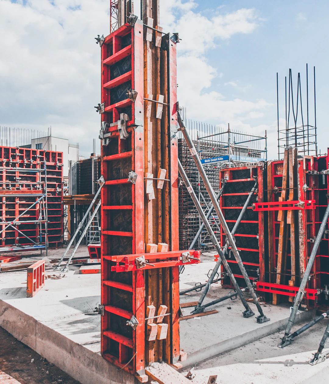 A inşaat sahası with multiple concrete formwork structures in place. Metal braces and supports are prominently visible, ensuring the stability of the forms. Concrete foundations and vertical columns are being prepared, with a mix of exposed rebar and newly laid concrete. The overall environment appears organized and active, reflecting ongoing building activities.