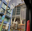 A tall modern building inşaat halinde is visible, surrounded by scaffolding and supported by large columns. In the foreground, there are road signs indicating directions, and a view of a city street with parked bicycles and cars. The scene has an urban atmosphere with a mix of inşaat malzemeleri and infrastructure.