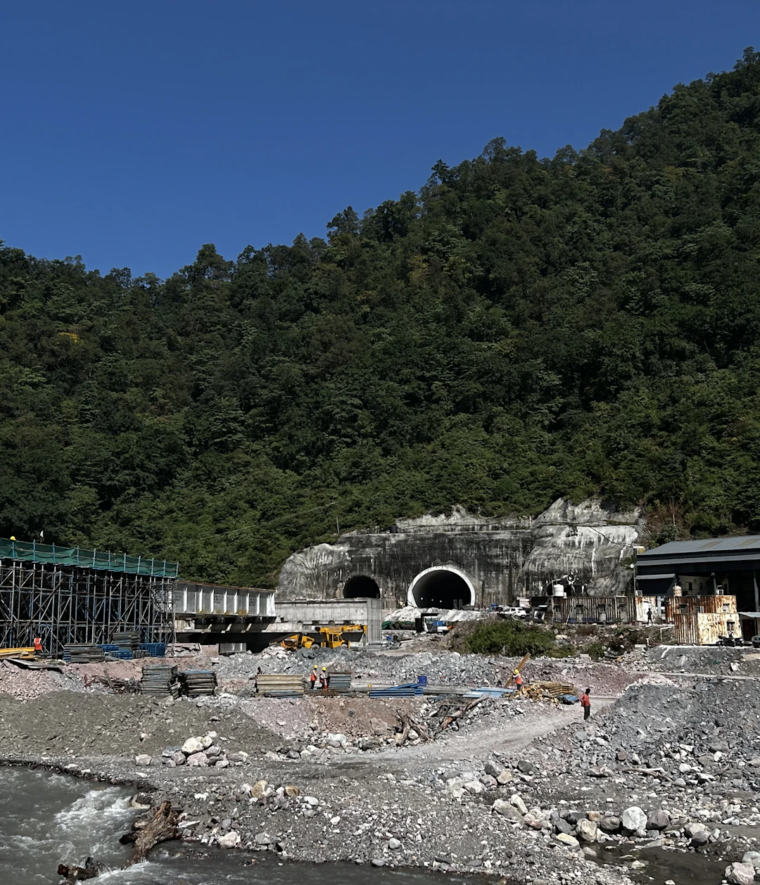 A inşaat sahası featuring a large tunnel entrance set against a forested hillside. There is a river flowing in the foreground with rocky banks. Various inşaat malzemeleri, scaffolding, and equipment, including vehicles and workers in safety gear, are scattered around the site. The sky is clear and blue, and the surrounding area is densely covered with trees.